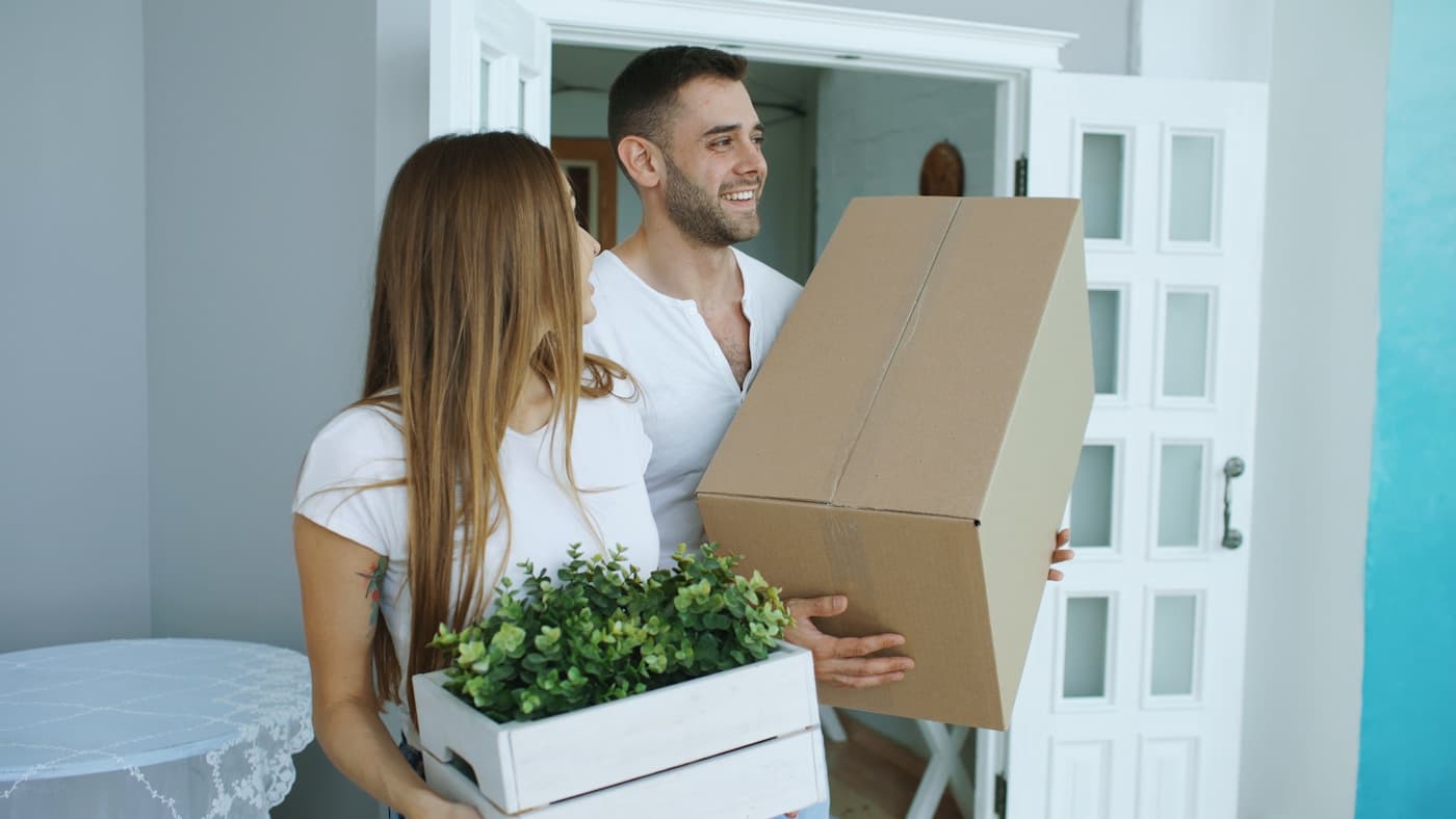 Two people carrying a cardboard carton through the front door of a home on moving day.